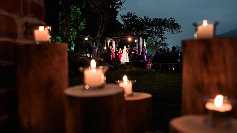 Casamento a céu aberto com velas na frente e noivos e convidados no fundo